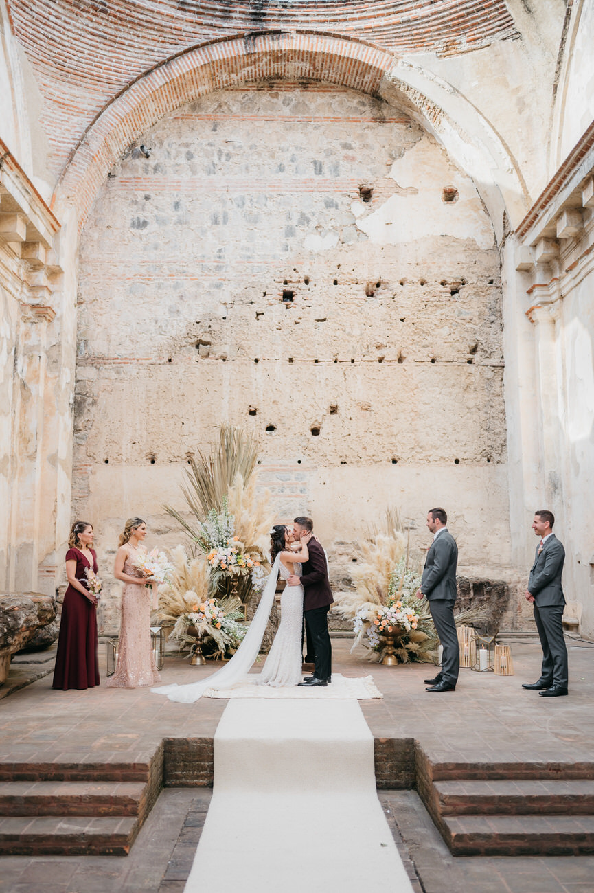 A couple shares their first kiss in the ruins of an old convent with a white aisle that leads up a few stairs to the altar.