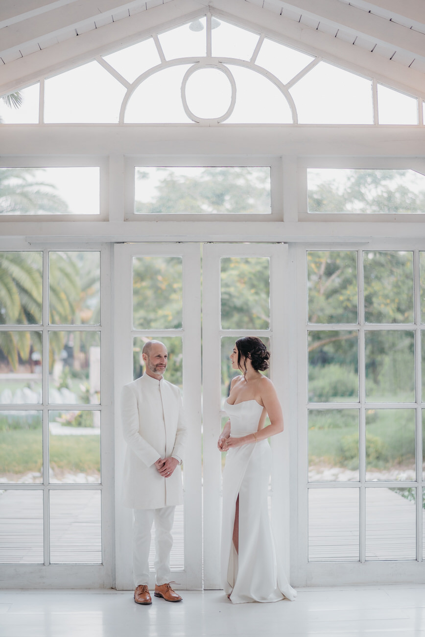 Bride and groom both dressed in white stand in a glass structure while looking in opposite directions