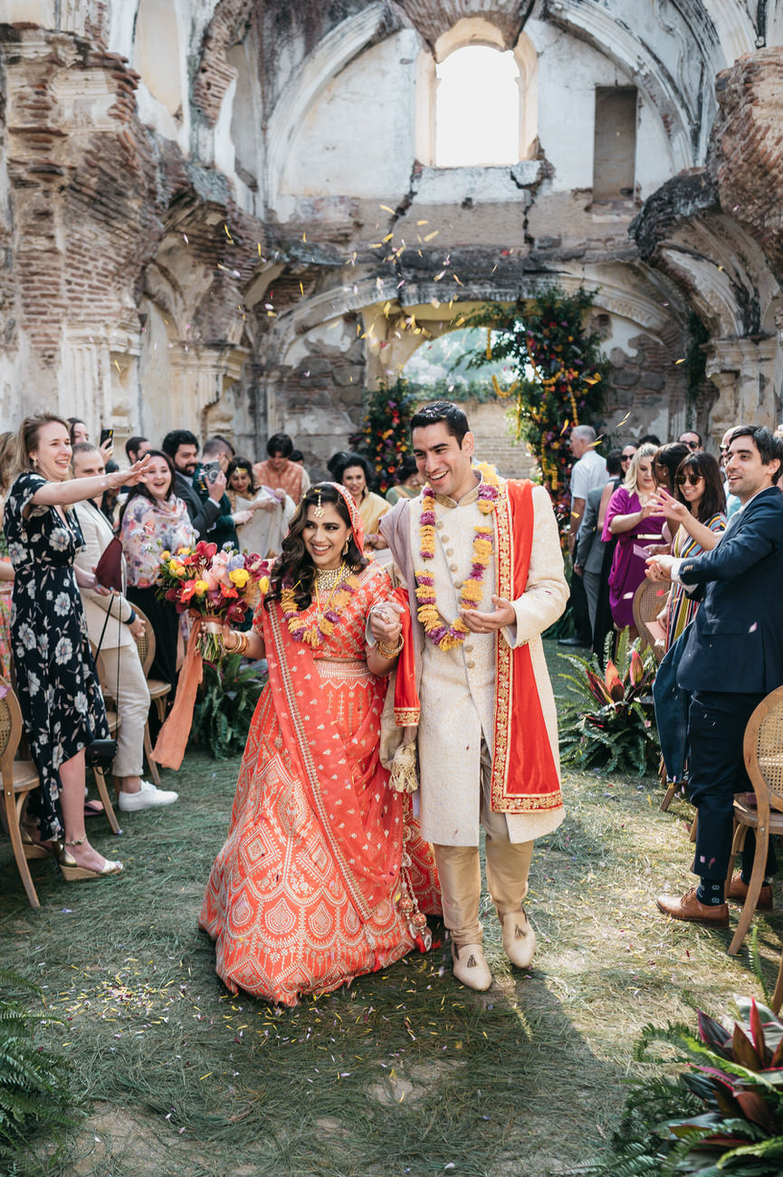 A wedding couple dressed in gold and red walk down the aisle while their guests throw red and yellow flower petals over their heads.