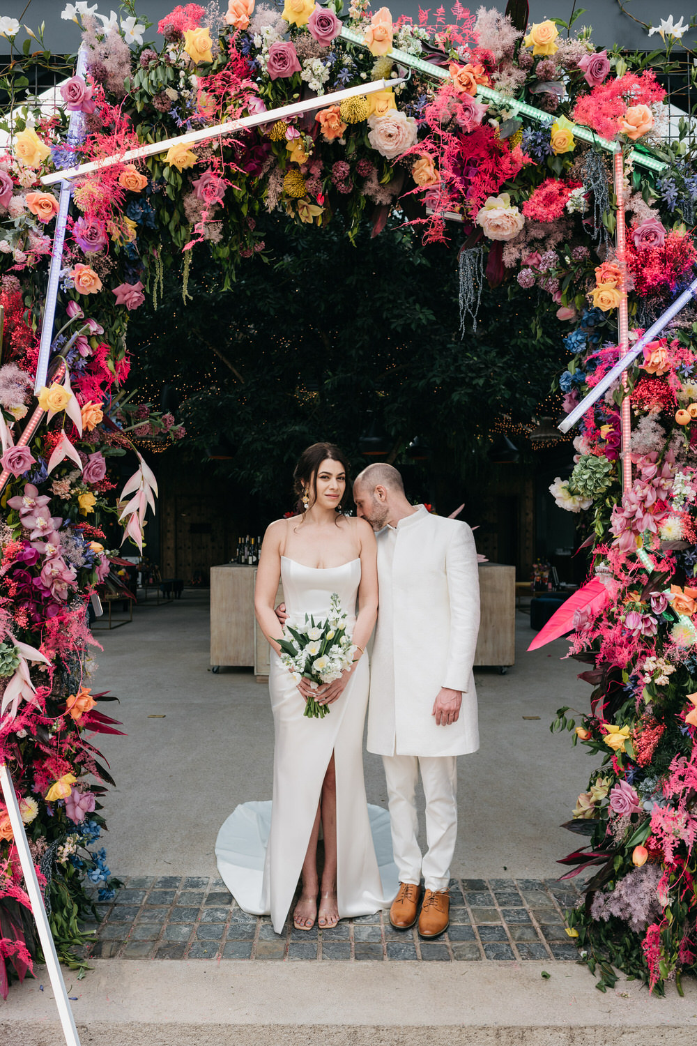 A wedding couple dressed in white stand in the middle of brightly colored floral arch with neon signs and textures decorating the arch.