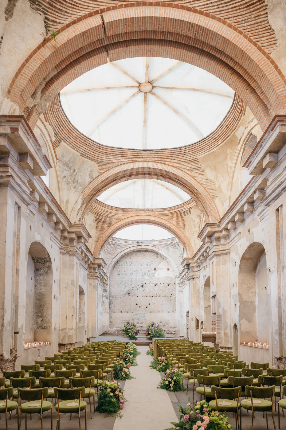 Ceremonia de boda en las Ruinas de Santa Clara con sillas de terciopelo verde oliva bordeando el pasillo de la ceremonia y una alfombra blanca larga que conduce al altar de bodas verde y rosa.