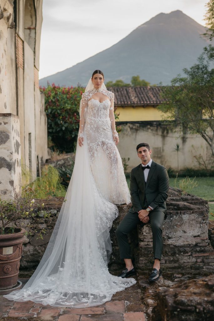 Bride stands on a ledge just next to the groom who is sitting next to her feet. There is a volcano in the background