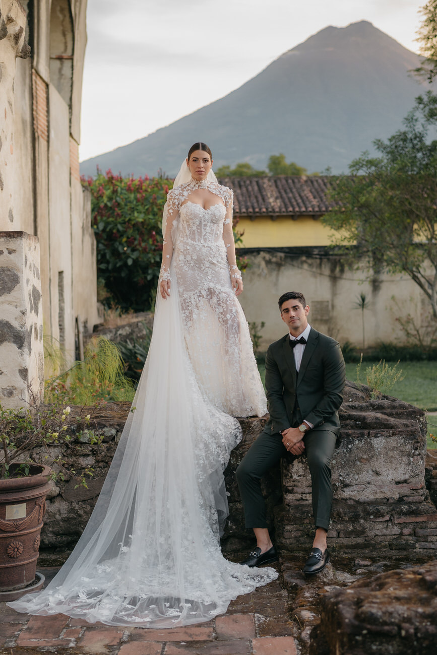 Bride stands on a ledge just next to the groom who is sitting next to her feet. There is a volcano in the background